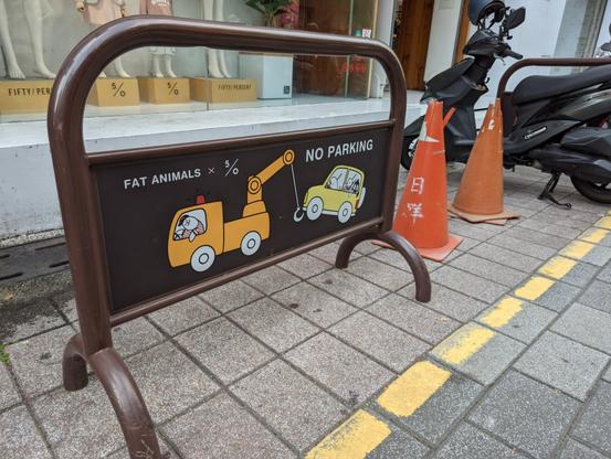 A knee-high No Parking barrier next to a small shop in Taiwan. Positioned on the sidewalk-less street it says in writing NO PARKING and shows a tow truck driven by a cat, and a towed car full of a happy Narwhale family in cute cartoon characters.