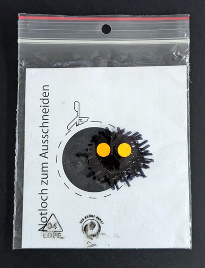 A piece of paper in a ziploc. It reads: Emergency hole to be cut out. Next to it a black circle with a cutting line. Drawn on top of the plastic with a sharpie: a little monster with yellow dots as eyes.