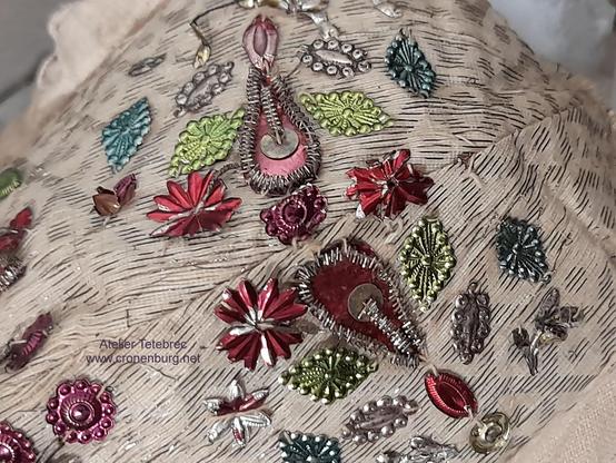 A white bonnet embroidered in colours with silver thread and floral embossed metallic elements. The elements were made in specialised factories. Because these were closed, restorers and conservators nowadays must build such elements from scrap by hand.
