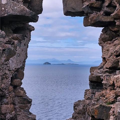 Photo through a hole in the stone wall of Duntulm Castle, Isle of Skye, with a view over the blue sea and the mountains of the Isle of Harris at the horizon.