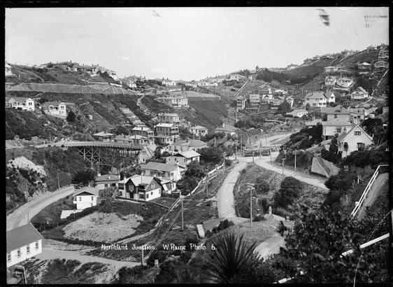 Black-and-white photo: Kelburn, Wellington. ca. 1920. Photographer William Hall Raine. Description: A view over the suburb of Kelburn, showing roads, houses and hills. In the middle distance is a tram moving away from the direction of the old wooden, single-laned viaduct, which was replaced in 1931. Citation: Radcliffe, Frederick George, 1863-1923: New Zealand post card negatives. Ref: 1/2-006699-G. Alexander Turnbull Library, Wellington, New Zealand. https://natlib.govt.nz/records/23033231