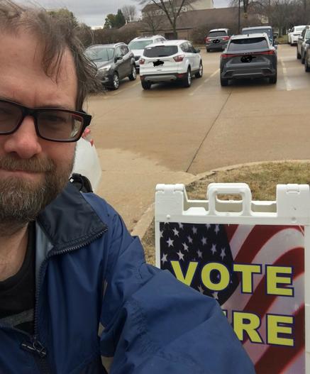 A selfie by scottmiller42 standing in front of a sign that has a USA flag and says "Vote Here".