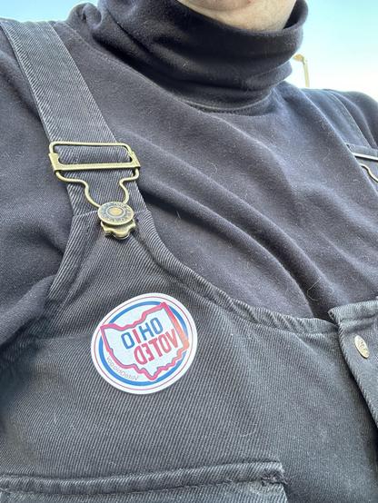 An “OHIO VOTED” sticker on the front right panel of black denim overalls (it’s actually a  button-down dress) worn over a black turtleneck. This may be an old goth who just voted against fascism.