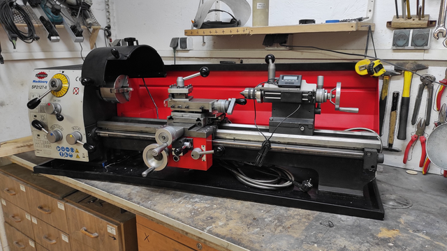 A mid-sized, benchtop lathe machine by SP2127-II, placed against a workshop wall with tools hanging in the background. The lathe has a red and black casing, with control levers and dials on the left side and the tool holder and chuck in the middle. The machine bed and rails are cleanly aligned, with a digital readout positioned above the chuck for precision. Various tools like screwdrivers, pliers, and measuring instruments are mounted on the wall, indicating an active workshop environment.