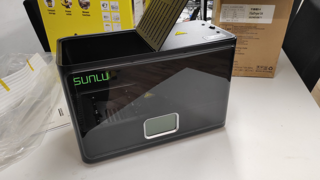 A black, boxy 3D printing filament dryer by SUNLU placed on a white tabletop. The front of the device has a clear viewing window, through which the interior mechanics, including the filament bed, can be seen. A small screen at the lower part of the device likely displays settings and temperatures. The dryer lid is open, showing the top of the filament compartment. Behind the device, some packaging and instruction materials are visible, along with a yellow box featuring icons of the dryer’s features.