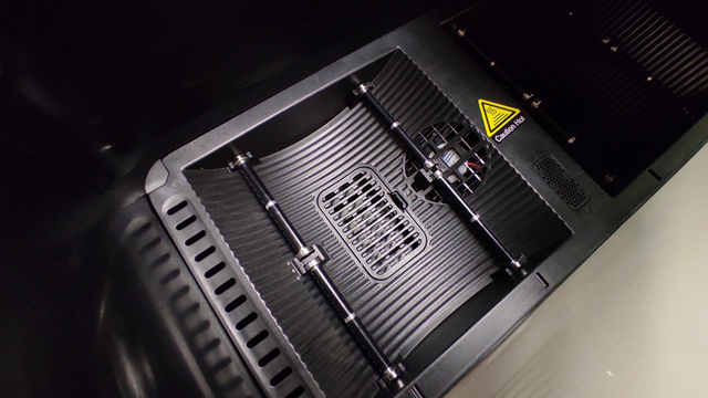 Close-up view of the interior of a 3D printing filament dryer. The image shows the filament bed with grooved, textured surfaces and two parallel rods with small clips to hold the filament spool. There is a circular fan vent near the top, and a caution symbol indicating "Hot" in yellow and black next to the vent. The components appear precisely arranged, highlighting the dryer’s structure for evenly heating and drying filament material for 3D printing.