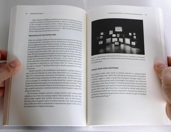 Hands hold a book open. The text header says "Repurposing and resurrecting media" and on the odd page there is a photo of sevelra television screens stack together and a text header "Analogue-digital media assemblages".