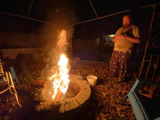 A man stands near a campfire at night, looking at his phone. The fire is glowing brightly, surrounded by a stone circle, with fallen leaves scattered on the ground. A few chairs are visible in the background.