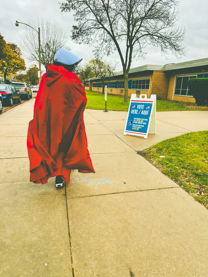Woman in a red gown and old fashioned white head covering from the handmaids tale is walking past a voting sign