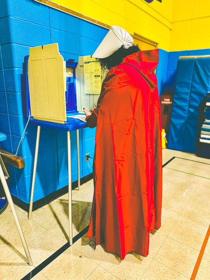 Woman in a red gown and old fashioned white head covering from the handmaids tale is standing at the voting booth voting