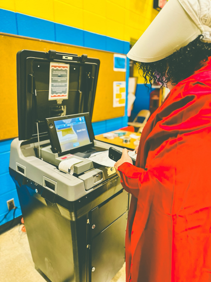 Woman in a red gown and old fashioned white head covering from the handmaids tale is sliding her ballot into the machine