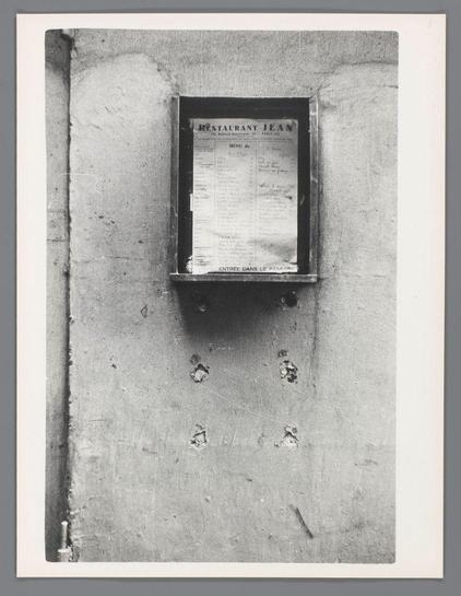 The image depicts a black and white photograph of an old-fashioned menu board mounted on the side of a weathered concrete wall. The menu appears to be for 'Restaurant Jean' located at 48 Boulevard Saint-Germain, Paris (partie). It features a list with French text indicating various dishes such as "ENTREE DANS LE PASSAGE," suggesting it's part of an entrance or passageway within the restaurant premises. Below this main menu is another section titled 'MENU du JOUR' which lists daily specials for April 1950, priced between FF3 and FF4 each. The paper shows signs of age with spots, tears at corners, and a creased top edge. To the left side where one might expect nails or screws to secure it in place are marks on the wall that suggest holes have been removed or filled over time.