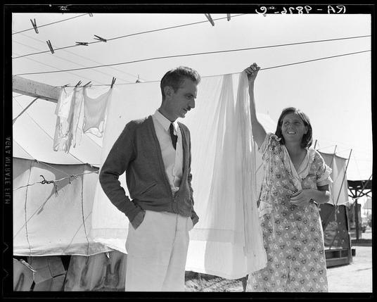 The image is a black and white photograph showing two individuals, likely in an outdoor setting with laundry hanging on the line behind them. The person to the left appears to be male and dressed in semi-formal attire consisting of a dark jacket, light-colored shirt, tie, and trousers; he has short hair and seems to be observing something out of frame with a contemplative expression.
The individual to the right is likely female based on her dress style, smiling broadly as she holds up a piece of white fabric. She wears a patterned knee-length skirt or dress with short sleeves, possibly floral in design, and stands confidently under what appears to be an awning supported by poles above their heads. The backdrop includes more hanging laundry, suggesting they might be at a campsite or temporary living area.
Additional context indicates that the photograph may have been taken during a time of displacement for migrants, as suggested by the mention of Kern migrant camp and Tom Collins' role in managing it.