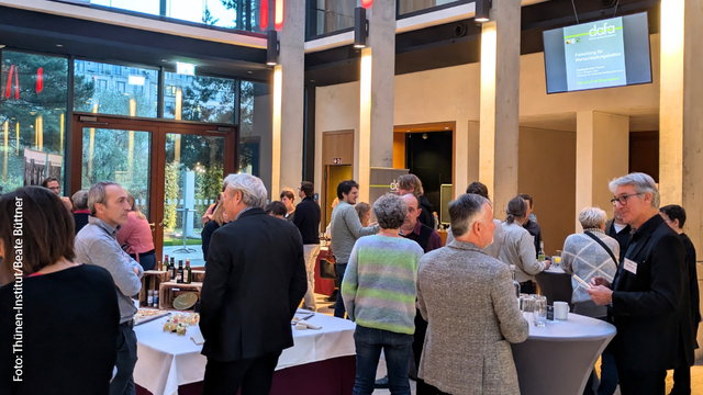 Das Bild zeigt eine Gruppe von Menschen, die sich in einem offenen Veranstaltungsraum zu einer Tagung versammelt haben. Die Teilnehmenden stehen an Stehtischen und im Raum verteilt, vertieft in Gespräche. Im Hintergrund sind große Fenster mit Blick auf Bäume und eine urbane Umgebung. Über einem der Tische hängt ein Bildschirm mit der Aufschrift „dafa - Forschung für Wertschöpfungsketten. Die Atmosphäre wirkt entspannt und professionell.