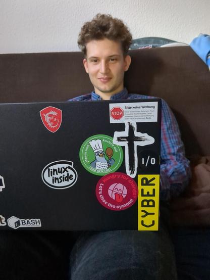 A guy sitting on a sofa working on a laptop computer. In the photo foreground you see its top cover with some stickers, most prominent a sticker from Coocook.org