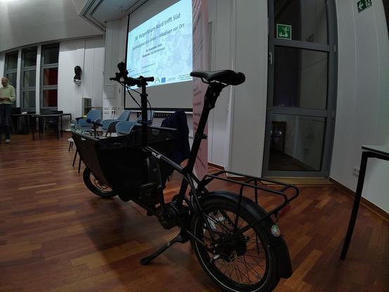 Ein schwarzes kompaktes Lastenrad mit Sticker der Kidical Mass Dortmund auf dem hinteren Schutzbleche. Auf einer Leinwand steht "38. Bürgerforum Nord trifft Süd"