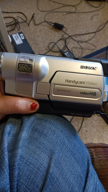 A silver mid-00s Sony HandycamVision video Hi8 camcorder than boasts a 560x zoom. Also a very chipped red painted thumbnail and my knee in blue jeans and a bunch of wires on the floor