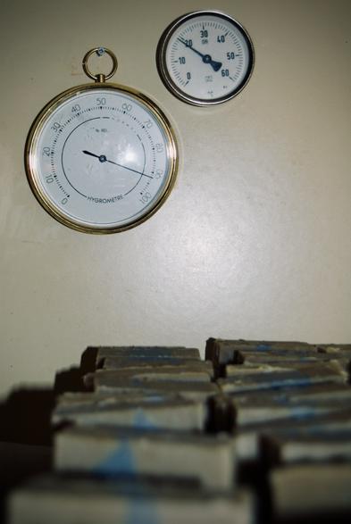 Sur le mur du séchoir à savon, il y a un thermomètre à aiguille (il fait 19°C) et un hygromètre (il fait 92% d'humidité). En première plan et en bas, il y a des savons verts et bleus qui sont rangés et en cure.