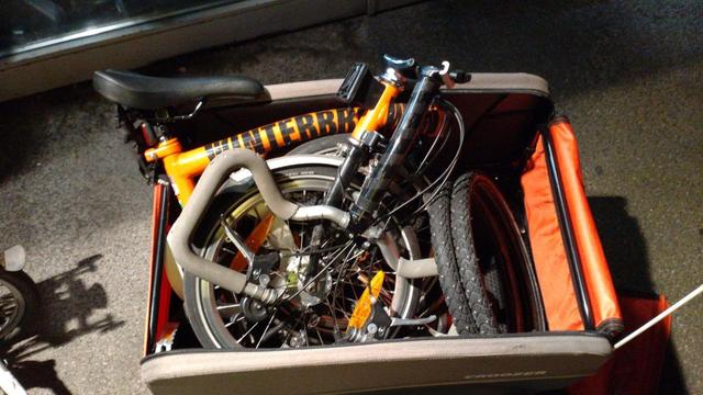 Ein zusammengefaltetes, orange-schwarzes Brompton-Faltrad mit schwarzem Schriftzug "WINTERBROMPTON" auf dem Hauptrohr liegt in einem orange-graubraunen Croozer Cargo Lastenanhänger mit geöffnetem Verdeck. Hinten im Lastenanhänger liegt noch ein paar Spikereifen. Ein Spike fehlt.