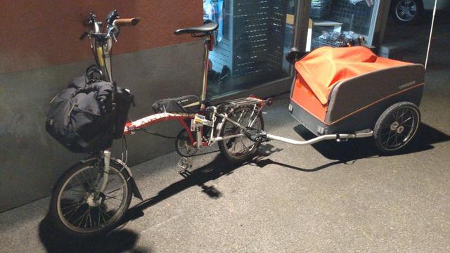Ein rote-weisses Brompton-Faltrad mit schwarzer Fronttasche und einem angehängtem orange-graubraunem Croozer Cargo Lastenanhänger mit nicht ganz zugehendem Verdeck und einem darunter herausguckenden Fahrradsattel steht vor einem erleuchteten Fahrradwerkstatt-Fenster.

Blick von schräg vorne, fast schon im Profil.