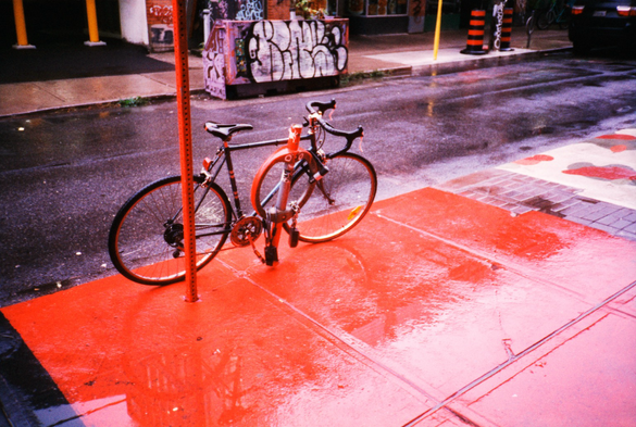 A bicycle is chained to a bicycle stand on a rainy sidewalk. The concrete of the sidewalk, the post of a traffic sign, an most of the bicycle stand are painted burnt umber reminiscent of an AMC from the seventies.