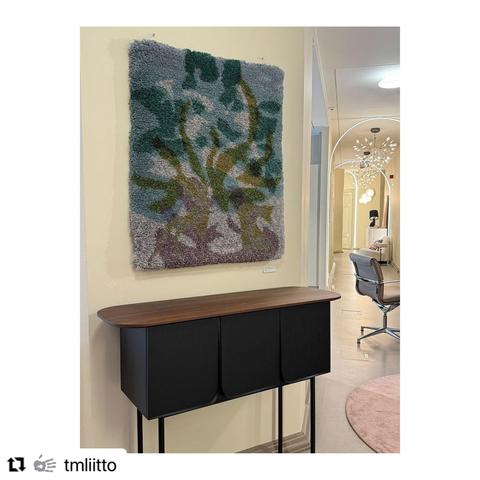 A screenshot of a vertical photograph on a white, square Instagram background. In the photo is an off-white interior space with ceiling lamps, a few different kinds of chairs, and in the foreground a thick-pile, wall-hanging rug on a cream-coloured wall over a dark, wooden sideboard on thin legs. The wall-hanging rug is known as a ryijy in Finland. It has a fuzzy texture that makes the rug's design look pixelated. The colours of the rug are pale teal, dark viridian green, grey yellow and purple, on grey background. The rug design is a stylised yellow flame overlapping with the teal smoke on the grey background. Where shapes overlap, the colours change.