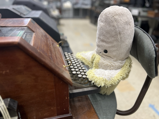 A stuffed banana sitting in a chair and using a teletypewriter