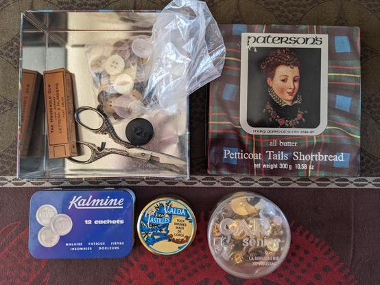 Une boîte carrée en fer-blanc, son couvercle posé à côté. Sur fond de tartan bleu et vert à filets rouges et jaunes, un portrait de la reine Marie Stuart, Paterson’s Petticoat Tails Shortbread. La boîte contient un sachet de boutons, une boîte d’étiquettes à vêtement, une paire de ciseaux guillochés. En dessous, une boîte de cachets Kalmine bleue et blanche, une petite boîte de pastilles Valda, un boîte en plastique transparent CAR Sénior, La Réglisserie à Moussac.