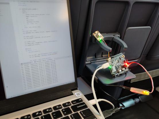 The back of two seats in a train. The drinks holder is flipped down and holds a SCD40 breakout, a Adafriit Metro RP2040, and a Raspberry Pi debug probe. On the left there is a Laptop screen showing some Rust code and terminal output.