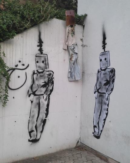 Deux silhouettes stylisées de pendus, la même masquée, peinte sur un mur. Entre les deux un tissus maculé de rouge est accroché.