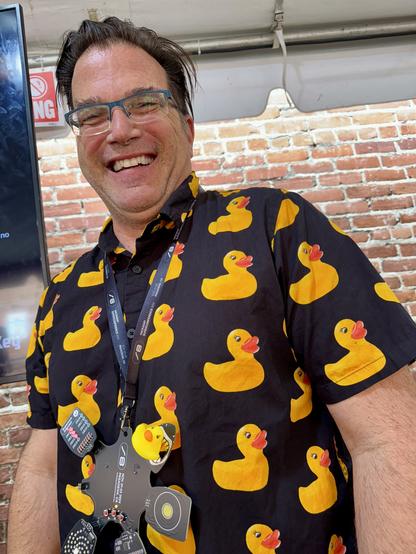 Man wearing a short-sleeve black shirt with yellow rubber duckies print, also wearing the Hackaday Supercon badge with the rubber duck SAO