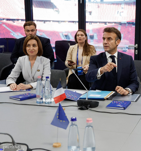 Foto de Macron y Maia Sandu poniendo caritas de q mierda es esto?? sentados a la mesa de la Comunidad Política Europea en Budapesta. Sobre la mesa, entre banderitas de sus países y de la Union Europea, hay banderas europeas y botellitas de AGUA.