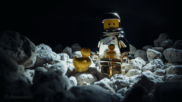 A golden Lego Classic Spaceman minifigure standing amongst small white pebble stones in hard light from above. On the figure’s chest there is a reflection of a Lego duck sitting on a pebble in front if it.