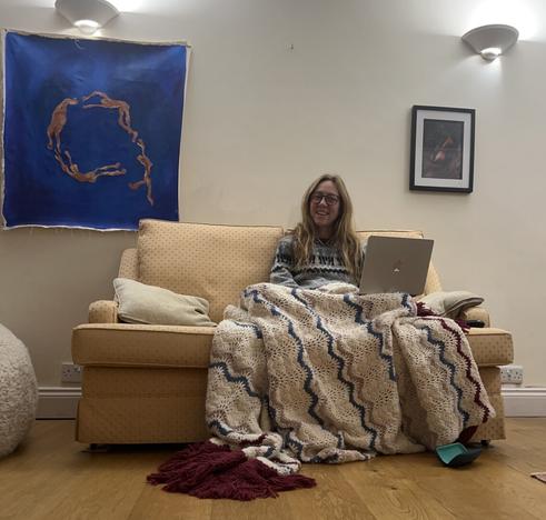 Smiling woman sitting on a peach sofa, typing on her laptop (on mastodon or Pixelfed of course!) she’s draped in a hand-crocheted ‘Afghan’ blanket made by her grandmother and her daughter’s art hangs above her - a beautiful blue painting of orange figures swimming or flying in a circle.