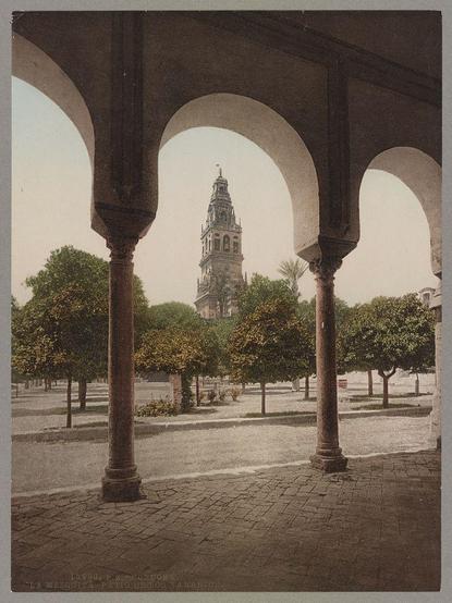 The image depicts an archway with three prominent columns leading the viewer's gaze through to a courtyard in the background. The architecture suggests a historical or religious significance, possibly from Spain given the mention of Córdoba and La Mezquita (a famous mosque). Through the archways, one can see neatly arranged trees lining paths within a spacious square-like area. A tall bell tower rises above this scene, further indicating that the location could be near a cathedral or significant religious site.

The sepia tone suggests that it is an old photograph, likely taken during the late 19th to early 20th century when such prints were popular for capturing architectural details and scenic views. The image serves as both artistic representation and historical documentation of this place in Córdoba, Spain.