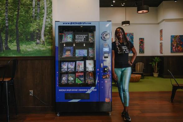Isis Asare, a Black person with long dreds, wearing a black t-shirt and blue pants, smiling and leaning against a blue vending machine stocked with books.