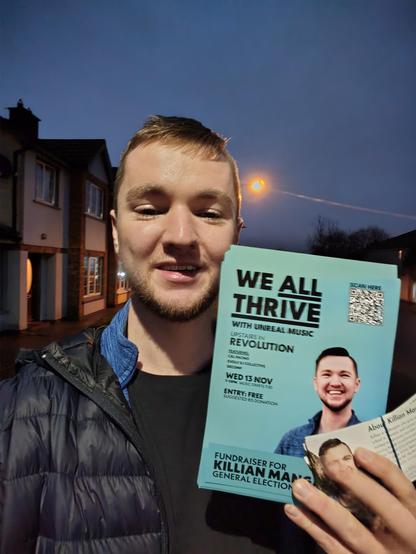 A selfie of Killian holding his We All Thrive with Unreal Music Fundraising Event leaflets and his campaign leaflets, with a dark sky behind him.