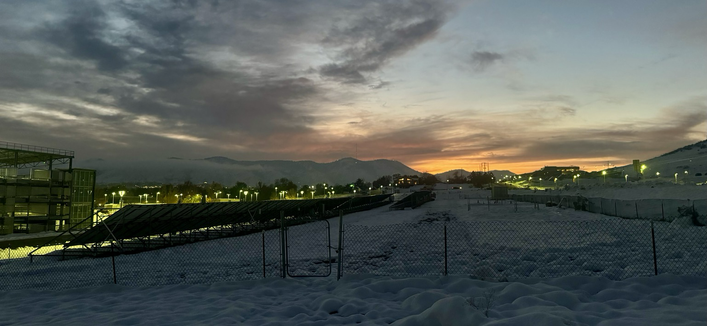 Snowy field with solar panels and city light on the left. Mountains and sunset in the background.