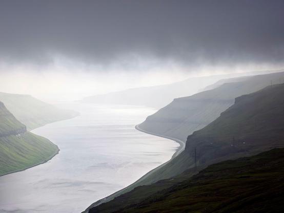 Under a dark foggy sky the water surface of a swinging fjord is reflecting the almost silvery sun light.