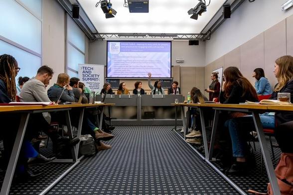 roundtable with 5 panelists seated at the front in front of conference banner and screen, facing an audience seated behind desks