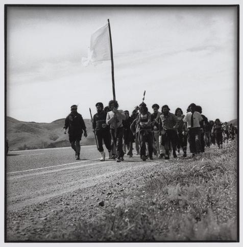 The photograph is a black and white image depicting a large group of people walking along the side of a road. The setting appears to be rural, with hills visible in the background under an overcast sky. At least 16 individuals are shown, many wearing casual or work-related attire such as jackets, shirts, pants, boots, gloves, hats, and various types of shoes. A few people can also be seen carrying items like a flag on a pole, a walking stick, or backpacks.

The group seems to have a cohesive purpose for their walk; they are all moving in the same direction along the road's shoulder with an organized formation. The presence of a single large white flag being carried by one person suggests that this could be part of some form of demonstration, march, religious pilgrimage, or cultural event.