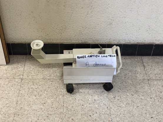 Un vieil appareil avec des roulettes en plastique est posé au sol. Une affichette dit : « Rover martien low tech — À donner ».