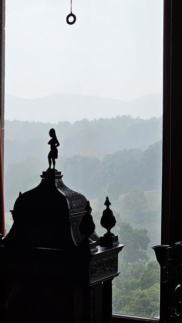 View out vertical window to four mountain ridges, each becoming more faint and gray until reaching smooth gray sky. at the top is a hanging pull loop for a window blind.

In the foreground toward the left, in silhouette, is an ornate clock with a female figure at the top.