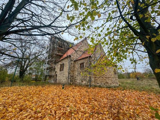 Kleine romanische Dorfkirche mit Gerüst hinter einer Herbstlaubwiese
