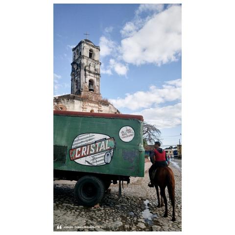 On the cobbled streets of Trinidad in Cuba, a worn vintage truck with a faded Cristal beer logo rests beneath the afternoon sun. A lone rider on horseback moves slowly past, merging seamlessly into the slow-paced rhythm of the old-world town. The bell tower of an old church stands watch, weathered yet proud, hinting at the stories it has witnessed over centuries.