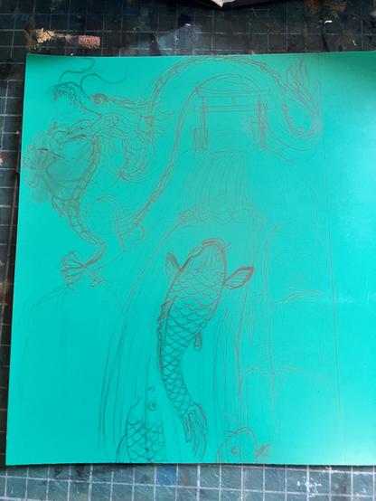 A pencil drawing on an uncarved green vinyl Lino block on a cutting mat with grid. The drawing shows three carp trying to climb a waterfall with gate at the top (like a Japanese torii gate). Across the top of the drawing is a Chinese style dragon. I’ve obviously erased, smudged and redrawn the front claw.
