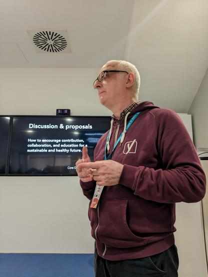 Andrea Fercia, in a Yoast hoodie, in front of one of his slides which reads "Discussions and proposals: how to encourage contribution, collaboration and education for a sustainable and healthy future"