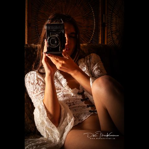 A woman in a lace dress lounges on a couch, a vintage camera aimed at the viewer.