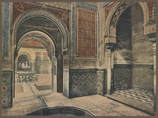 The image depicts an intricately designed room within the Alhambra, a historic palace complex in Granada, Spain. The architecture is characterized by vibrant and detailed geometric tile work on walls and floors, as well as ornate archways leading into other parts of the structure. A central fountain can be seen through one of these openings, surrounded by what appears to be stone animal statues or sculptures at its base. The room's ceiling features Islamic calligraphy art in deep blue hues, creating a sense of cultural richness and historical significance. Sunlight filters through an opening above, casting dynamic shadows on the tiled surfaces below. This photograph is part of a collection documenting various aspects of the Alhambra, including halls, courtyards, fountains, and palatial architecture from Granada.