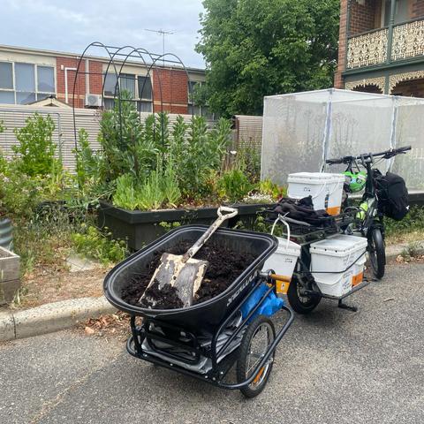 An e-bike towed wheelbarrow, filled with compost, parked in front of slightly overgrown raised beds